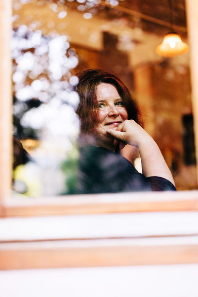 Portrait Heidi Sekulla blickt über die Schulter aus einem Fenster in die Kamera